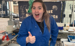 Zara, a white appearing female with brown/blonde hair smiling at the camera with her thumb up.